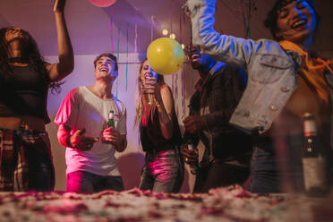 Young men and women having fun at a colorful house party with decorations and confetti all around. Friends dancing and celebrating holding drinks at a house party. - JLPSF01096