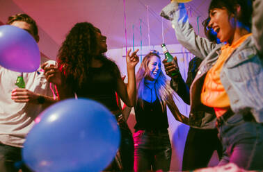 Junge Männer und Frauen, die sich auf einer bunten Hausparty mit Dekorationen und Konfetti amüsieren. Freunde tanzen und feiern mit Getränken auf einer Hausparty. - JLPSF01097