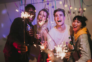 Friends celebrating with fire sparkles and drinks at a house party. Young men and women having fun at a colorful house party with decorations and confetti all around. - JLPSF01100