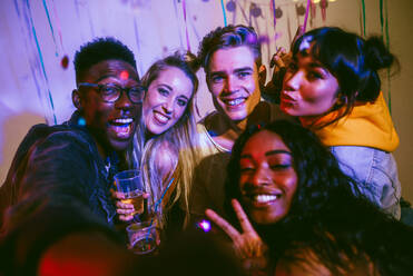Friends posing for a selfie holding drinks and showing victory signs at a house party. Young men and women having fun at a colorful house party. - JLPSF01103