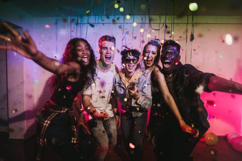 Young men and women having fun at a colorful house party with decorations and confetti all around. Friends celebrating and shouting in joy posing for a photograph at a house party. - JLPSF01113