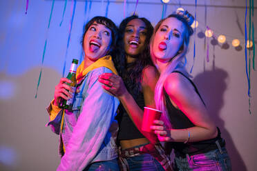 Drei junge Frauen stehen zusammen und schneiden Grimassen, während sie auf einer Hausparty Getränke in der Hand halten. Freunde, die für ein Foto posieren, strecken ihre Zungen heraus und haben Spaß auf einer bunten Hausparty. - JLPSF01115