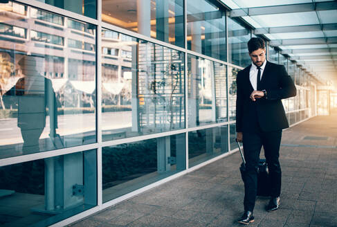 Geschäftsmann, der einen Koffer trägt und auf seine Uhr schaut, während er im Flughafen spazieren geht. Geschäftsreisender, der die Zeit auf seiner Armbanduhr überprüft. - JLPSF01123