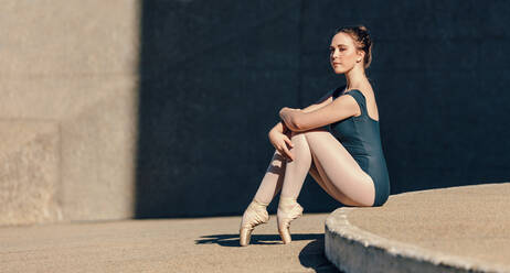 Balletttänzerin sitzt anmutig im Freien. Balletttänzerin sitzt in Spitzenschuhen und stützt ihre Zehen auf den Boden. - JLPSF01129