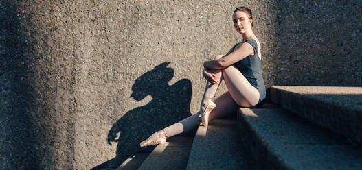 Balletttänzerin sitzt im Freien und nimmt eine Pose ein. Balletttänzerin sitzt auf einer Treppe und streckt ihre Knöchel in Spitzenschuhen. - JLPSF01138