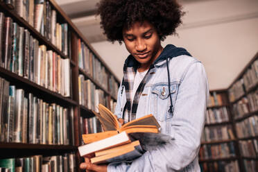 Afrikanischer männlicher Student in der Bibliothek mit vielen Büchern stehend, Universitätsstudent auf der Suche nach Studienunterlagen. - JLPSF01143