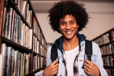 Junger afrikanischer Mann mit Rucksack, lächelnd und in die Kamera blickend, steht in der Bibliothek. - JLPSF01144