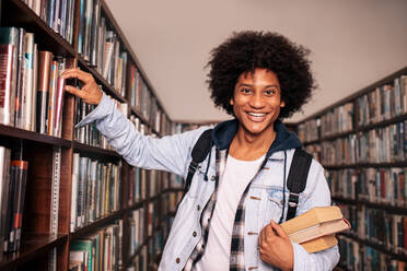 Lächelnder junger schwarzer Mann, der ein Buch aus dem Regal nimmt. Universitätsstudent, der ein Buch aus dem Regal nimmt und in die Kamera schaut. - JLPSF01145