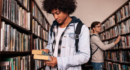 Junger afrikanischer Student, der ein Buch in der Universitätsbibliothek betrachtet. College-Studenten auf der Suche nach einem Buch in der Bibliothek. - JLPSF01146