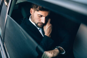Young businessman talking on the mobile phone and looking at the watch while sitting on back seat of car. Caucasian business executive checking time and making phone call in car. - JLPSF01150