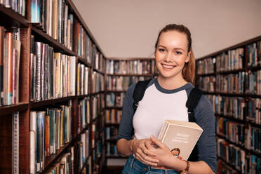 Junge kaukasische Studentin steht mit einem Buch in der Bibliothek. College-Student trägt ein Buch in der Bibliothek. - JLPSF01151