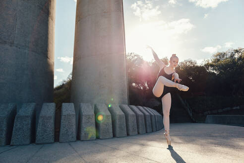 Balletttänzerin beim Üben von Tanzschritten im Freien mit Sonne im Hintergrund. Tänzerin steht auf einem Zeh in Spitzenschuhen und übt ihre Tanzschritte. - JLPSF01154