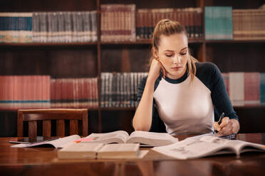 Studentin, die sich in der Bibliothek auf die Prüfung vorbereitet. Studentin, die in der Bibliothek sitzend Notizen aus einem Buch macht. - JLPSF01159