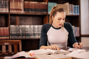 Weibliche Studentin macht sich Notizen aus einem Buch in der Bibliothek. Junge kaukasische Studentin sitzt am Tisch und erledigt Aufgaben in der College-Bibliothek. - JLPSF01161