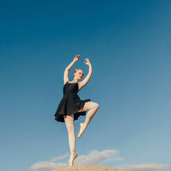 Balletttänzerin beim Üben von Tanzschritten im Freien. Tänzerin steht auf einem Zeh in Spitzenschuhen auf einem Felsen mit blauem Himmel im Hintergrund. - JLPSF01170