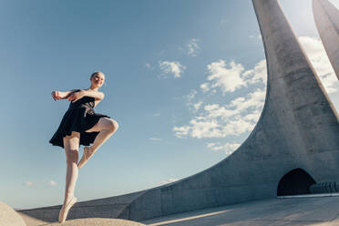 Ballet dancer practicing dance moves outdoors with blue sky and monument in the background. Female dancer balancing on one toe in pointe shoes on a rock. - JLPSF01172
