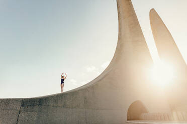 Ballet artist practicing dance moves outdoors. Female dancer standing on a monument practicing dance steps with sun in the backdrop. - JLPSF01189