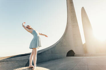 Balletttänzerin übt Tanzschritte im Freien. Tänzerin steht an einem Denkmal und übt Tanzschritte mit Sonne im Hintergrund. - JLPSF01192