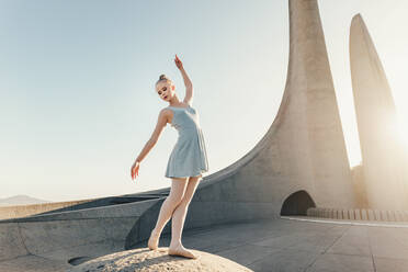 Ballettkünstlerin übt Tanzschritte im Freien. Tänzerin steht an einem Denkmal und übt Tanzschritte mit Sonne im Hintergrund. - JLPSF01193