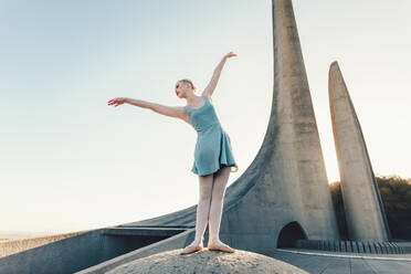 Ballettkünstlerin übt Tanzschritte im Freien. Tänzerin steht an einem Denkmal und übt Tanzschritte mit Sonne im Hintergrund. - JLPSF01194