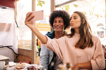 Junge Frau macht Selfie mit afrikanischem Mann in Cafeteria. Junges Paar schaut auf Smartphone in Café. - JLPSF01196