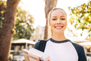 Porträt einer schönen Studentin, die im Freien lächelt. Junge Studentin steht mit einem Buch auf dem College-Campus. - JLPSF01210