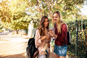 Schöne College-Studenten schauen sich Bilder auf dem Handy an. Junge Frauen mit Buch schauen auf das Smartphone. - JLPSF01217