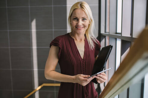 Porträt einer Frau, die mit einem digitalen Tablet die Treppe im Büro hinaufgeht. Weibliche Führungskraft bei der Arbeit in einem modernen Büro. - JLPSF01234