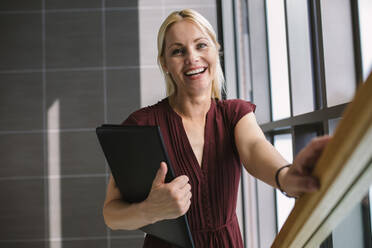 Geschäftsfrau, die die Treppe in einem Bürogebäude hinaufgeht. Kaukasische Frau hält eine Akte und lächelt. - JLPSF01235