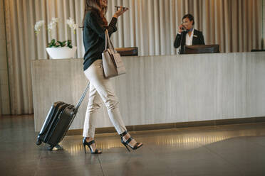 Geschäftsfrau geht mit Gepäck vor der Hotelrezeption und telefoniert. Weiblicher Gast kommt mit Koffer im Hotel an. - JLPSF01259
