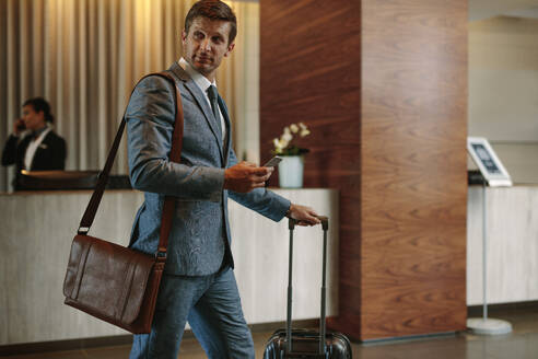 Geschäftsmann geht mit Handy und Gepäck durch einen Hotelflur. Männlicher Geschäftsreisender kommt in seinem Hotel an. - JLPSF01265