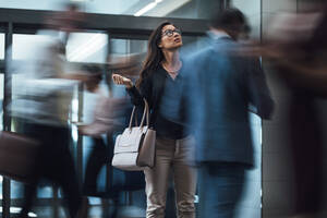 Geschäftsfrau, die steht und nach oben schaut, während sie auf jemanden wartet, während Menschen in der Lobby eilen, Bewegungsunschärfe-Effekt. - JLPSF01266