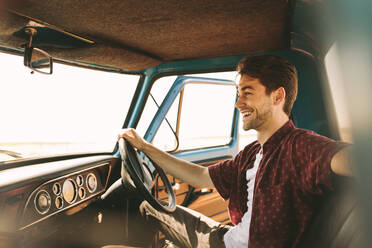 Ein lächelnder Mann sitzt auf dem Fahrersitz eines alten Autos. Ein Mann sitzt in seinem Oldtimer und hält das Lenkrad bei geöffneter Tür. - JLPSF01272