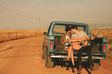 Ein Ehepaar sitzt auf dem Rücksitz ihres Pickups und genießt die Fahrt auf dem Land. Der Mann spielt Gitarre, während die Frau eine Flasche mit einem Getränk in der Hand hält. - JLPSF01294