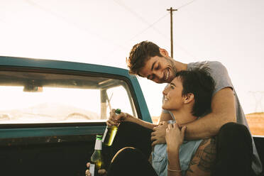 Smiling couple sitting in the back of their pickup truck enjoying the road trip in country side. Man and woman holding a bottle of drink enjoying the road trip. - JLPSF01298