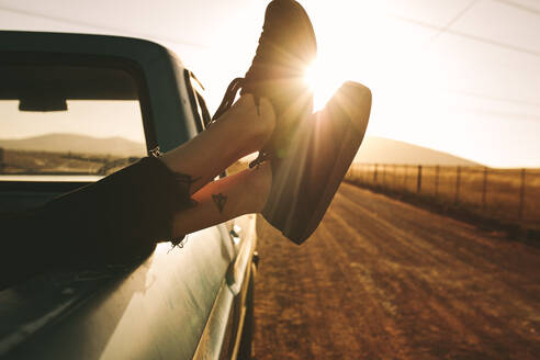 Nahaufnahme der Beine einer Frau, die sich auf der Ladefläche eines Pick-up-Trucks auf einer Landstraße ausruht, mit der Sonne im Hintergrund. - JLPSF01300