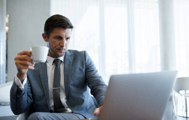 Business executive working on laptop and having coffee at office. Caucasian businessman looking at laptop with coffee in hand. - JLPSF01322