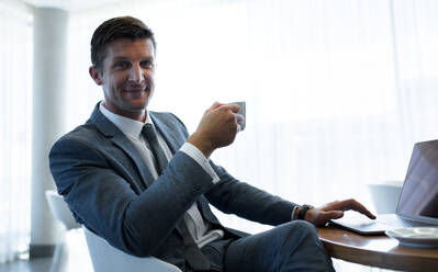 Gutaussehender Geschäftsmann sitzt an einem Tisch mit Laptop und trinkt Kaffee. Männliche Führungskraft macht eine Kaffeepause im Büro. - JLPSF01323