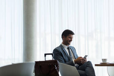 Business traveler sitting at airport waiting area reading text message on his mobile phone. Businessman waiting for flight at airport lounge. - JLPSF01329