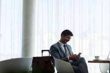 Glücklicher Geschäftsmann, der in der Flughafenlounge auf sein Mobiltelefon wartet. Geschäftsreisender mit Smartphone, der auf seinen Flug wartet. - JLPSF01330