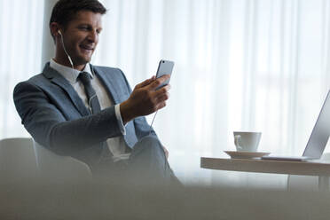 Businessman with smartphone having video call at office. Caucasian man in suit sitting at office lobby using mobile phone for making video call. - JLPSF01333