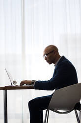 Seitenansicht eines Geschäftsmannes, der an einem Tisch sitzt und an einem Laptop im Büro arbeitet. Ein Unternehmer benutzt einen Laptop im Büro. - JLPSF01344