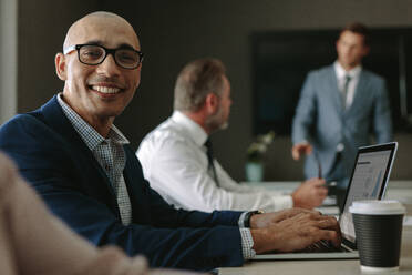 Lächelnder Geschäftsmann während einer Besprechung im Konferenzraum. Männliche Führungskraft sitzt im Büro im Vorstandszimmer und diskutiert mit Kollegen im Hintergrund. - JLPSF01354