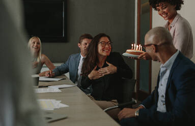 Office workers celebrating birthday of cheerful female colleague with cake in office. Surprise birthday celebration of a female associate during a staff meeting. - JLPSF01365