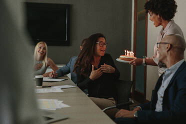 Office workers celebrating birthday of cheerful female colleague with cake in conference room. Coworkers congratulating female colleague on birthday and presenting cake with candles. - JLPSF01366