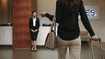 Rückansicht einer Geschäftsfrau, die mit ihrem Gepäck im Hotel ankommt. Im Hintergrund steht eine weibliche Concierge, um den Gast zu begrüßen. - JLPSF01369