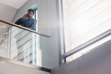 Geschäftsmann lehnt an einem Geländer in einem Bürogebäude und spricht mit seinem Smartphone. - JLPSF01399