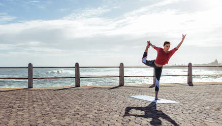 Woman stretching during yoga on road along the sea. Fit woman workout outdoors, doing Natarajasana, Dancer king pose on seaside promenade. - JLPSF01402