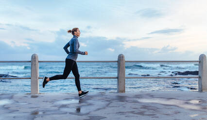 Seitenansicht von Fitness-Frau, die auf einer Straße am Meer läuft. Sportlerin, die auf der Strandpromenade trainiert. - JLPSF01418