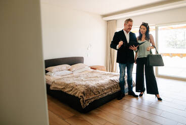 Male real estate agent showing apartment and discussing the contract and documents with a female customer. Realtor showing the contract papers to female client while standing inside the bedroom of a new house. - JLPSF01433